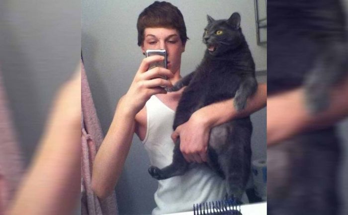 A large grey cat being held up for a selfie in the bathroom mirror and not looking happy about it 